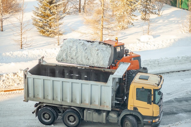 El gran tractor naranja limpia la nieve de la carretera y la carga en el camión. Limpieza y limpieza de caminos en la ciudad de nieve en invierno. Remoción de nieve después de nevadas y ventiscas.