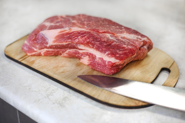 Gran trozo de carne fresca sobre una tabla de cortar de madera con un cuchillo de metal sobre una mesa de cocina ligera