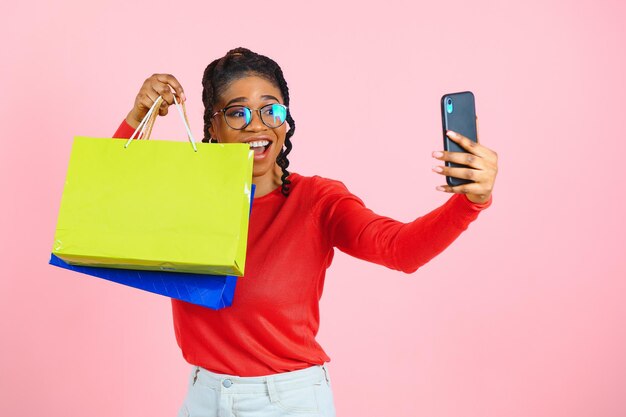 Gran venta Emocionada chica afro con bolsas de compras tocando gafas de sol mirando a la cámara sobre la pared rosa del estudio