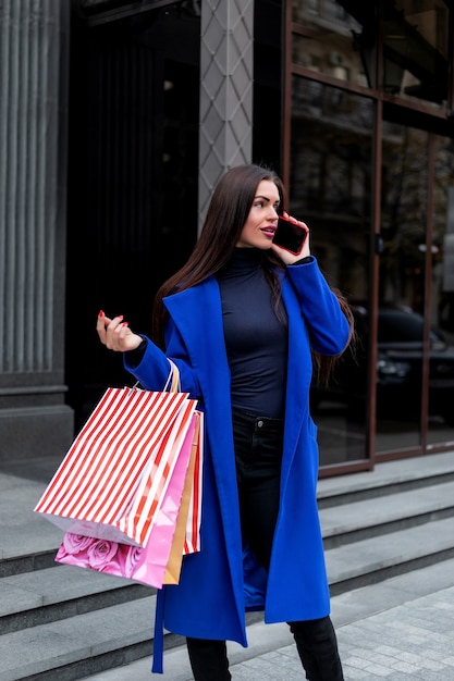 Gran venta. Mujer de moda joven hermosa con bolsas de la compra junto a la ventana de la tienda llamando a teléfonos inteligentes, tonos. mujer de compras, divertido, teléfono