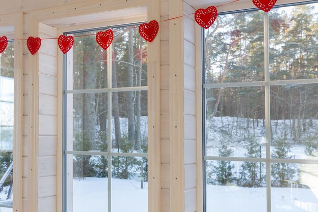 Una gran ventana de dormitorio blanco en una casa de madera decorada con guirnaldas románticas de corazones rojos en el Día de San Valentín. Luminoso interior de la habitación en una casa de madera con una gran ventana con vistas al invierno