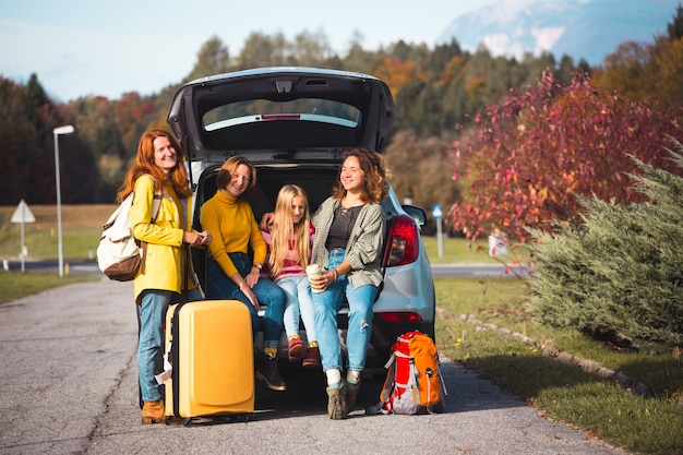 Foto gran viaje familiar: las niñas felices viajan en coche. mamá con hijas sentadas en el maletero