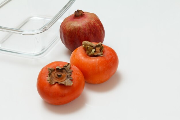 Granada madura fresca, frutas de caqui y un recipiente de vidrio sobre la superficie blanca