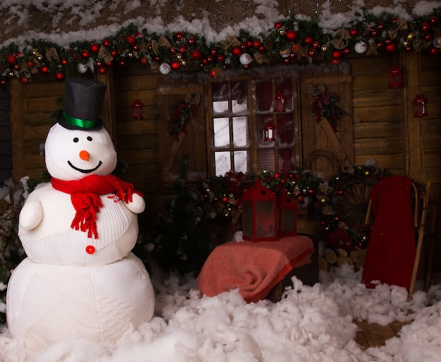 Grande boneco de neve de inverno com boné em pé na casa de madeira decorada com algodão neve no chão.