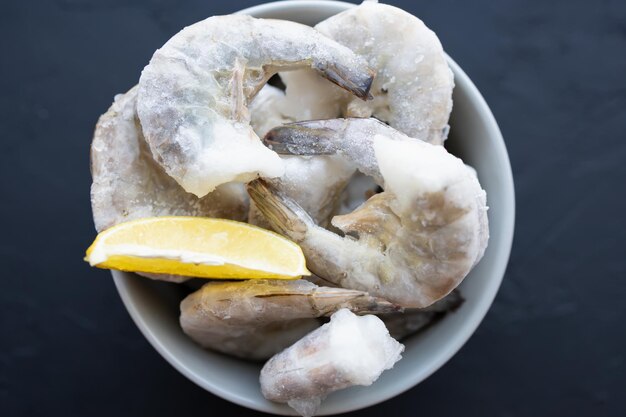 Grande camarão congelado em tigela cinzenta em fundo preto com limão gelado para cozinhar delicioso camarão-rei sem cabeça para preparação de refeições desfrute de frutos do mar naturais com baixo teor de gordura e alto teor de proteína foco suave