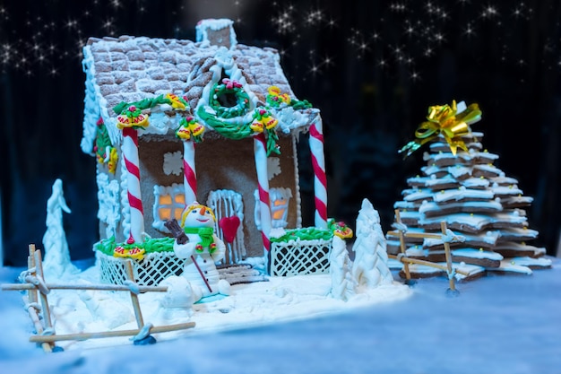 Grande casa de pão de mel caseiro coberta de neve, árvore de natal de pão de mel e um boneco de neve de mástique de açúcar à noite. maquete para ofertas sazonais e cartão postal de férias