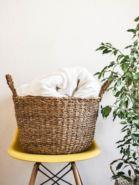 Foto grande cesta de vime com um cobertor branco em uma cadeira amarela e planta de casa contra uma parede branca.