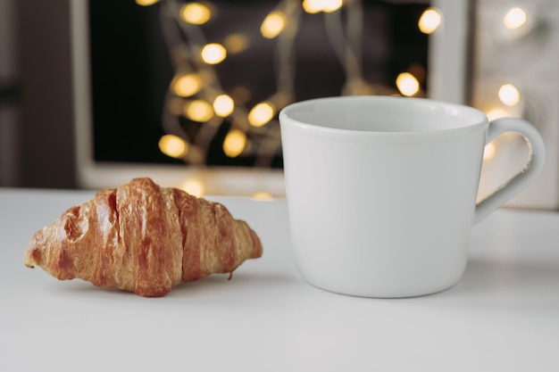 Foto grande croissant em um prato com uma grande caneca branca de café no fundo de uma guirlanda fechada