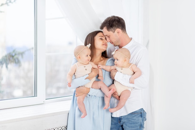 Grande família feliz com gêmeos mãe e pai segurando duas gêmeas recém-nascidas em um terno de algodão na janela da casa abraçando e beijando-os maternidade e paternidade com dois filhos