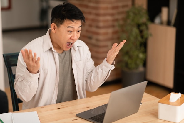 Grande oferta online Homem asiático emocional usando laptop e gritando lendo notícias sentado em casa e espalhando as mãos