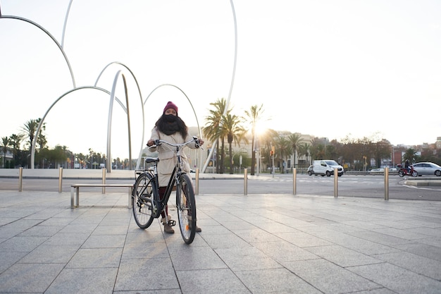 Grande plano de uma jovem afro-americana em pé com uma bicicleta contra o sol em uma praça da cidade moderna.