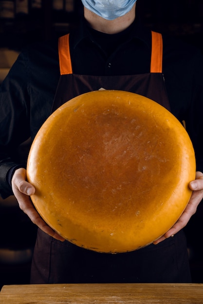Grande roda de queijo amarelo nas mãos. vendedor com máscara para proteção contra o coronavírus covid-19. segurando o queijo redondo.