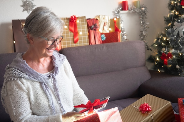 Grande sorriso de uma mulher sênior, recebendo um novo tablet como presente de Natal. Cabelo grisalho e óculos. Muitos presentes ao redor dela