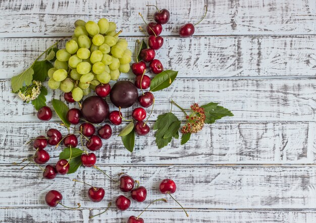 Grandes cerejas doces suculentas, ameixas e uvas, folhagens e flores em um fundo de madeira