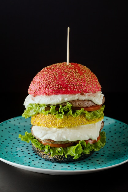 grandes hambúrgueres com cores diferentes pão pães em vermelho, amarelo e preto com creme de parmesão e tomate, salada em cima da mesa.