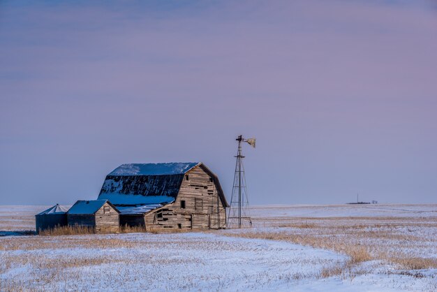 Granero vintage, contenedores y molino de viento rodeado de nieve bajo un cielo rosado al atardecer en Saskatchewan, Canadá