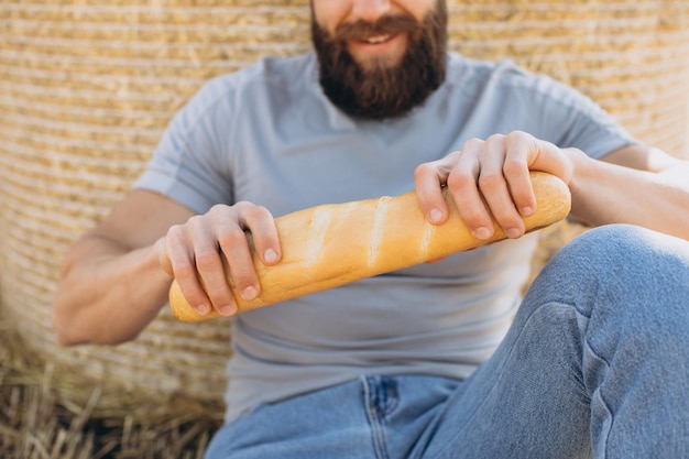 Granjero barbudo feliz en jeans sosteniendo pan fragante fresco y sentado cerca de la paca alrededor del campo de sol soleado