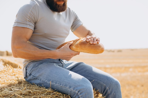 Granjero barbudo en jeans sosteniendo pan fresco y fragante lo rompe por la mitad sentado encima de una paca alrededor de un campo soleado