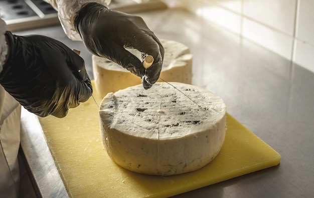 Un granjero con guantes negros corta una cabeza de queso gorgonzola picante con moho azul con una cortadora en pedazos