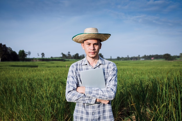 Granjero de hombre asiático de pie con la mano que sostiene la computadora portátil en la granja de arroz verde, computadora portátil