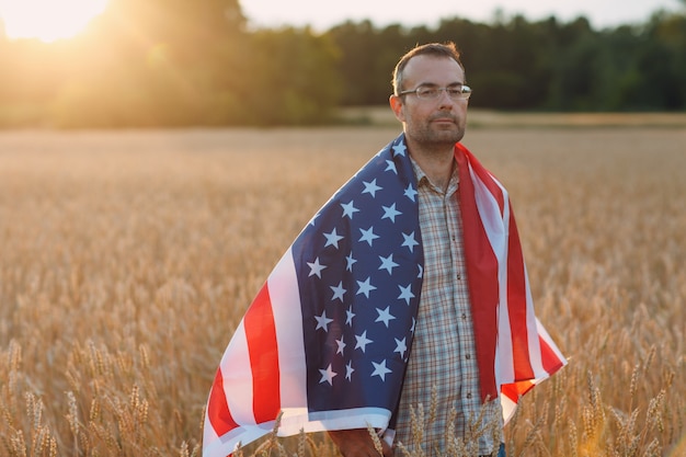 Granjero de hombre que cubre la bandera americana en el campo de trigo al atardecer agricultura y cosecha agrícola