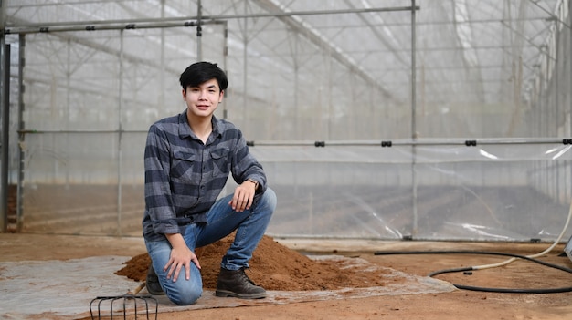 Granjero joven guapo en camisa a cuadros y jeans sentado cerca de un montón de tierra para plantar en invernadero.