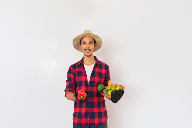 Granjero negro con sombrero y guantes sosteniendo una canasta de verduras (zanahoria, limón, tomate, chayote y remolacha) aislado en fondo blanco.