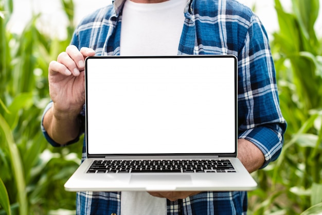 Granjero que se coloca en un campo que sostiene la computadora portátil abierta. Maqueta de pantalla blanca
