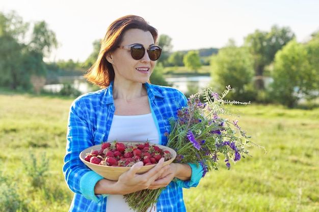 Granjero de sexo femenino con tazón de fuente de fresas recién cosechadas y ramo de flores silvestres. Fondo de naturaleza, paisaje rural, prado verde, estilo campestre