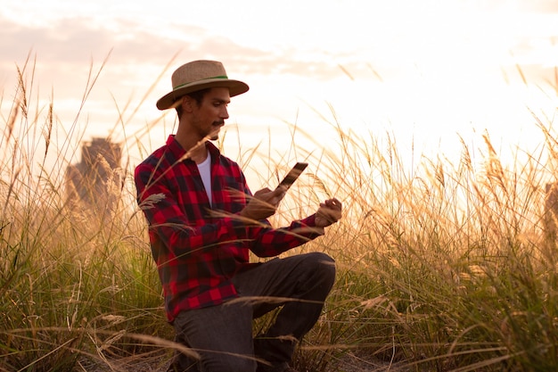 Granjero con sombrero y tableta móvil analizando la plantación al atardecer.