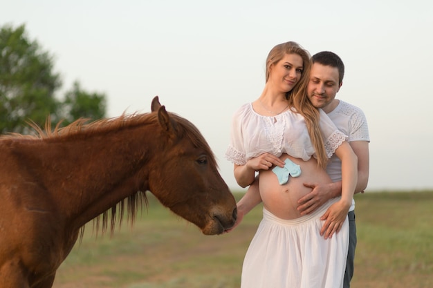 Un granjero con su esposa embarazada al atardecer en su granja. Posando con un caballo.