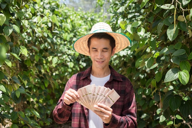 Un granjero tailandés con una camisa roja sostiene dinero de billetes tailandeses en una granja