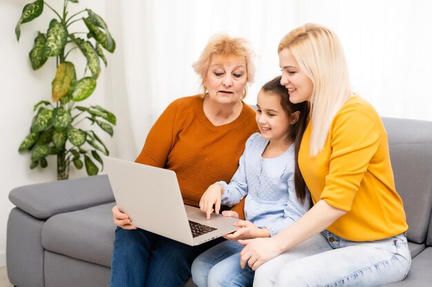 Granny, su hija y su nieta están usando una computadora portátil y sonriendo mientras están sentadas en el sofá en casa.