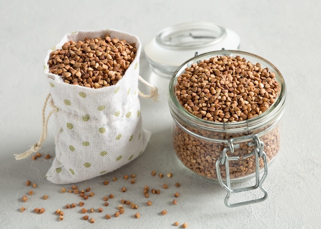 Grano de trigo sarraceno crudo en un frasco de vidrio y cereales en una bolsa de lona de cerca sobre un fondo de hormigón gris.