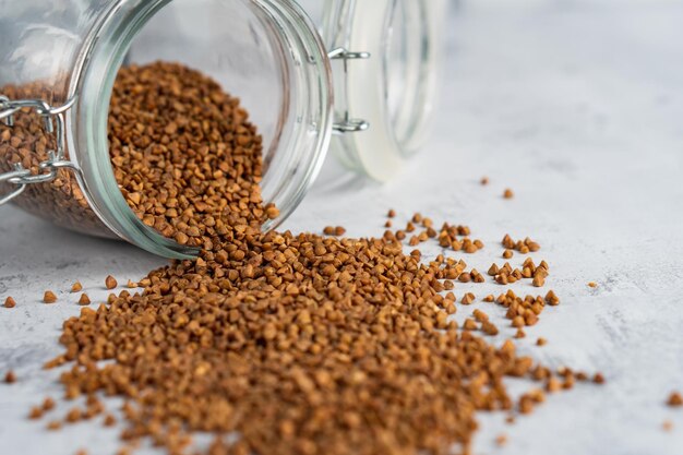 Grano de trigo sarraceno de primer plano vertido de un frasco de vidrio sobre una mesa gris Enfoque selectivo