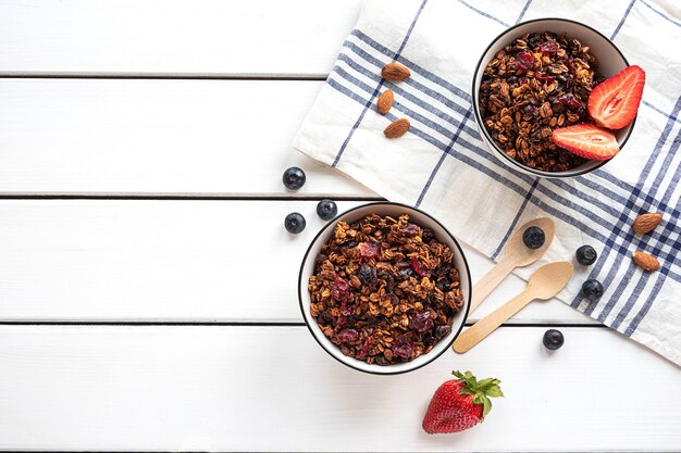 Granola assada caseira em duas tigelas com frutas vermelhas, amêndoas, guardanapo e colheres ecológicas na mesa branca, vista de cima