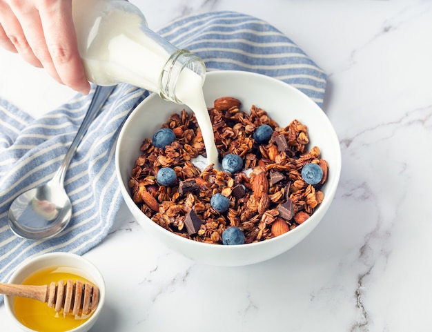 Granola caseira, muesli com pedaços de chocolate escuro, nozes, mirtilos e despejando leite em uma tigela na superfície de mármore branco. café da manhã saudável.