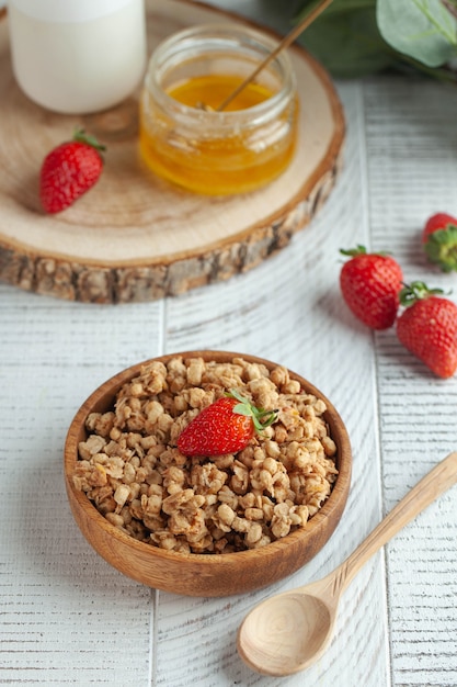 Granola casera en un cuenco de madera con fresas y miel sobre una superficie de madera blanca