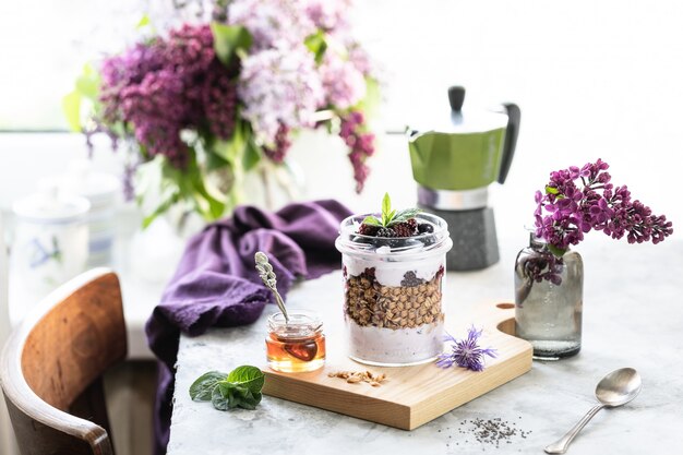 Granola casera en un frasco con yogur griego y moras y frambuesas sobre la mesa con lila.