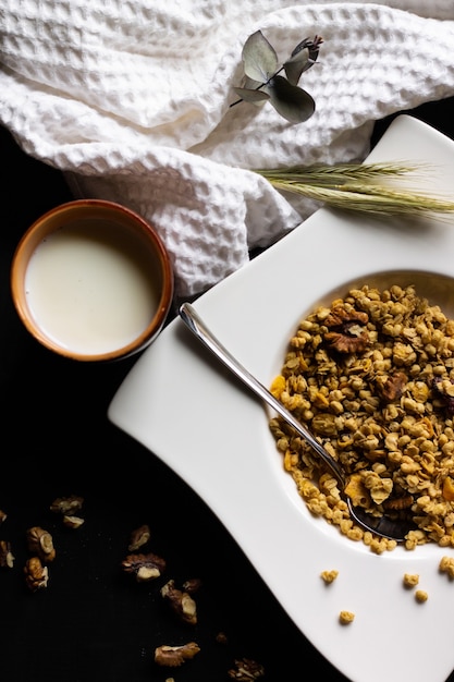 Granola de cereais em um prato branco com uma colher e um copo de barro com leite sobre uma mesa de madeira preta. Vertical. Vista do topo. Foco seletivo