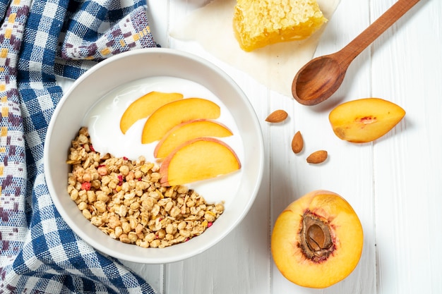 Granola de trigo branco com iogurte desnatado em uma tigela branca em uma composição com uma colher, favos de mel e pêssego em fundo branco de madeira.