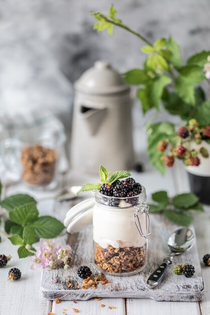 Granola hecha a mano con yogurt natural blanco con moras en un frasco de vidrio transparente, flores y hojas sobre un fondo blanco de madera.