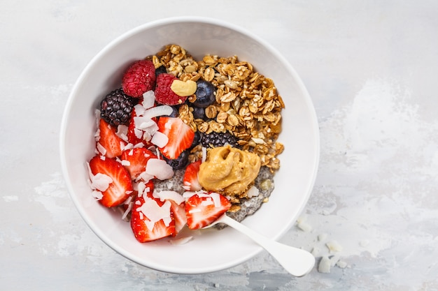 Granola con leche, chia, bayas y mantequilla de maní en un tazón blanco, vista superior.