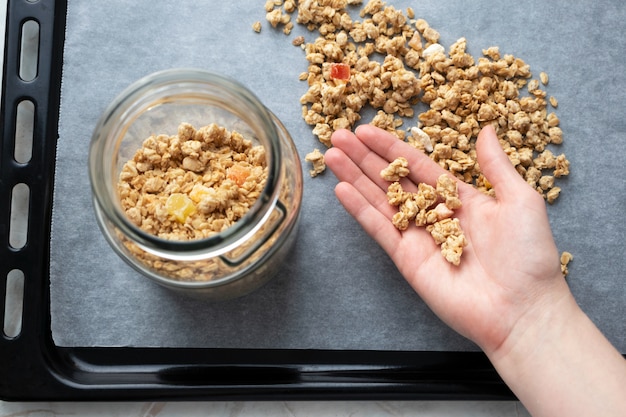 Granola en mano femenina y frasco de vidrio sobre papel pergamino en una bandeja para hornear, vista superior. Desayuno casero hecho en casa