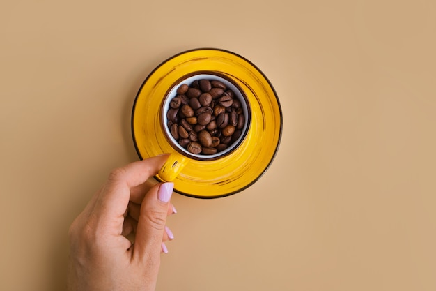 Granos de café arábica en una taza de diseño amarillo brillante con platillo en mano femenina sobre un papel beige pastel