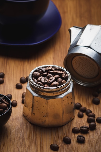 Granos de café en una cafetera géiser sobre una mesa de madera con una taza en un plato.