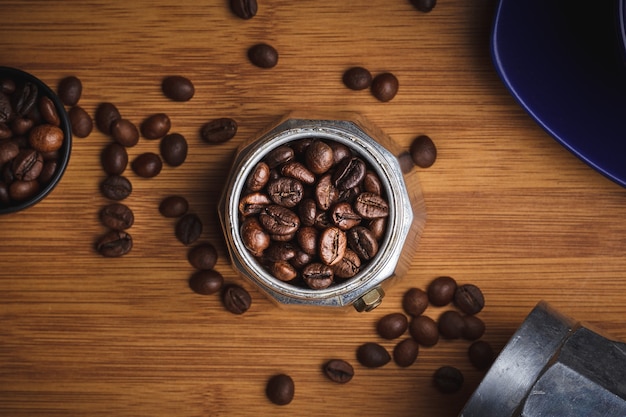 Granos de café en una cafetera géiser sobre una mesa de madera con una taza en un plato.