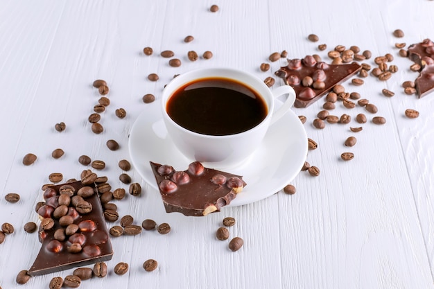 Granos de café dispersados, una taza y chocolate negro en una tabla de madera blanca. Copia espacio