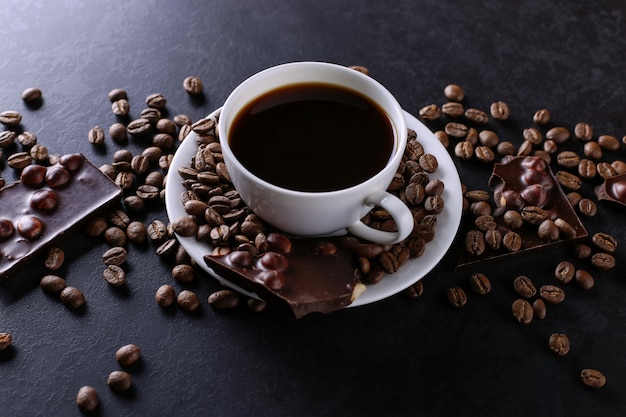 Granos de café dispersos, una taza y chocolate negro sobre una mesa de piedra negra. Copia espacio