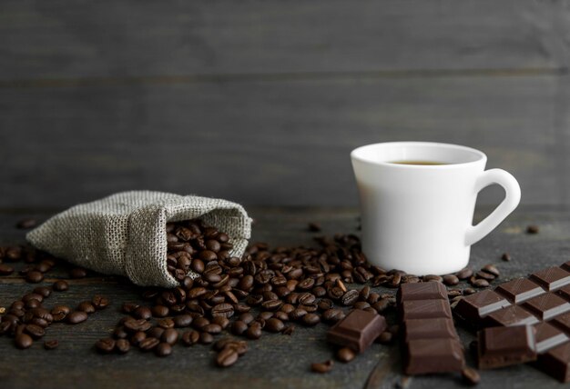 Granos de café esparcidos de una bolsa de lino sobre una mesa de madera Taza de café y barra de chocolate negro